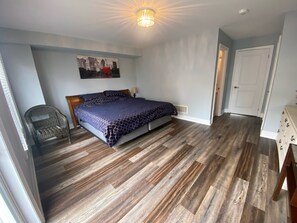 A bedroom with a bed, wooden flooring, a chair, a door, and a wall-mounted picture.