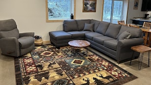 Una sala de estar con un sofá seccional gris, un sillón gris, una mesa de centro de madera y un tapete estampado.
