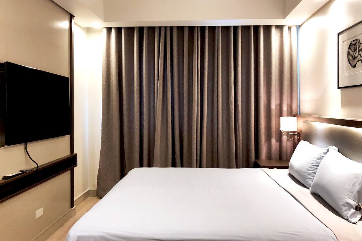 A hotel room with a bed, a flat-screen TV mounted on the wall, a nightstand with a lamp, and a large window with curtains.