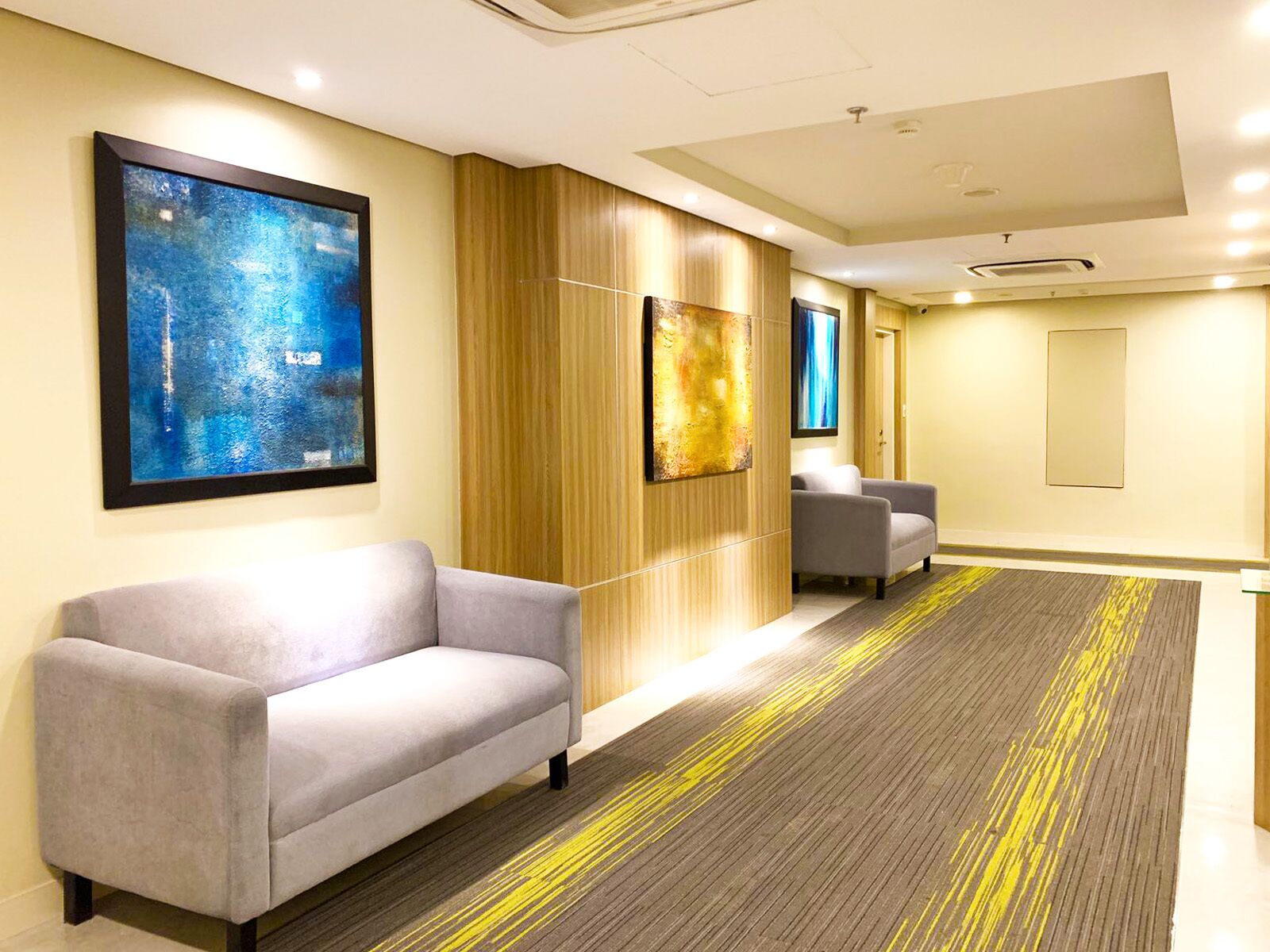 A hotel lobby with a grey sofa, a wooden panel wall, and two abstract paintings.