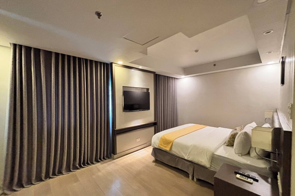 A modern hotel room with a large bed, a flat-screen TV mounted on the wall, and a wooden floor.