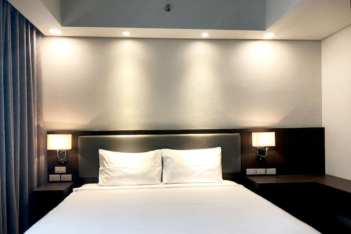 A hotel room with a large bed, two bedside lamps, a dark headboard, and a ceiling light.