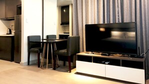 Un espacio habitable moderno con televisor de pantalla plana sobre un mueble blanco, una mesa de comedor con sillas y una zona de cocina con refrigerador y hervidor.