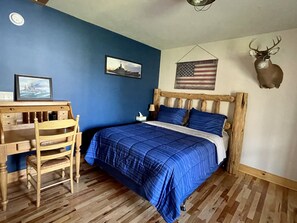 Une chambre à coucher avec un lit en bois, une literie bleue, un bureau avec une chaise, un tableau encadré et une tête de cerf fixée au mur.