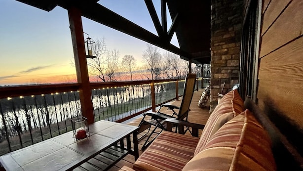 Une terrasse en bois avec un banc et une table, donnant sur une étendue d’eau bordée d’arbres, avec un coucher de soleil en toile de fond.