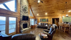 L’intérieur chaleureux d’un chalet avec une cheminée en pierre, des fauteuils en cuir, un coin repas et de grandes fenêtres offrant une vue imprenable.