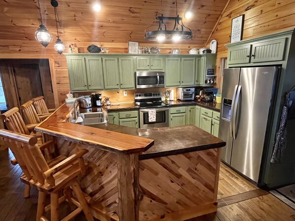 A wooden cabin-style kitchen with green cabinets, a central island with bar stools, stainless steel appliances, and hanging pendant lights. - Secluded lakeside cabin, with a salt water pool & hot tub, on 25 acres! (Austin)
