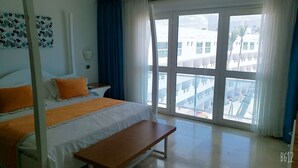 A hotel room with a bed, a bench, a nightstand, and a window with a view of buildings. - Playa Resort hamaca  (Boca Chica)