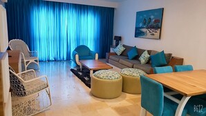 A living room with a sofa, coffee table, ottomans, and chairs. There is a painting on the wall and a window with blue curtains. - Playa Resort hamaca  (Boca Chica)