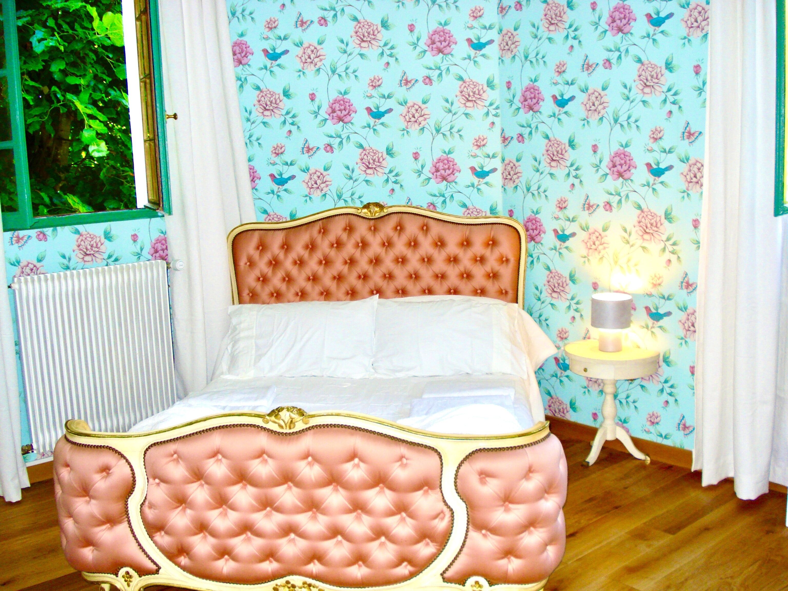 A bedroom with a tufted headboard, floral wallpaper, a bedside table with a lamp, and a window with curtains.