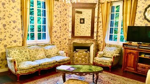 Un salon traditionnel avec un papier peint à motifs floraux, une cheminée, un canapé, un fauteuil, une table basse et un téléviseur.