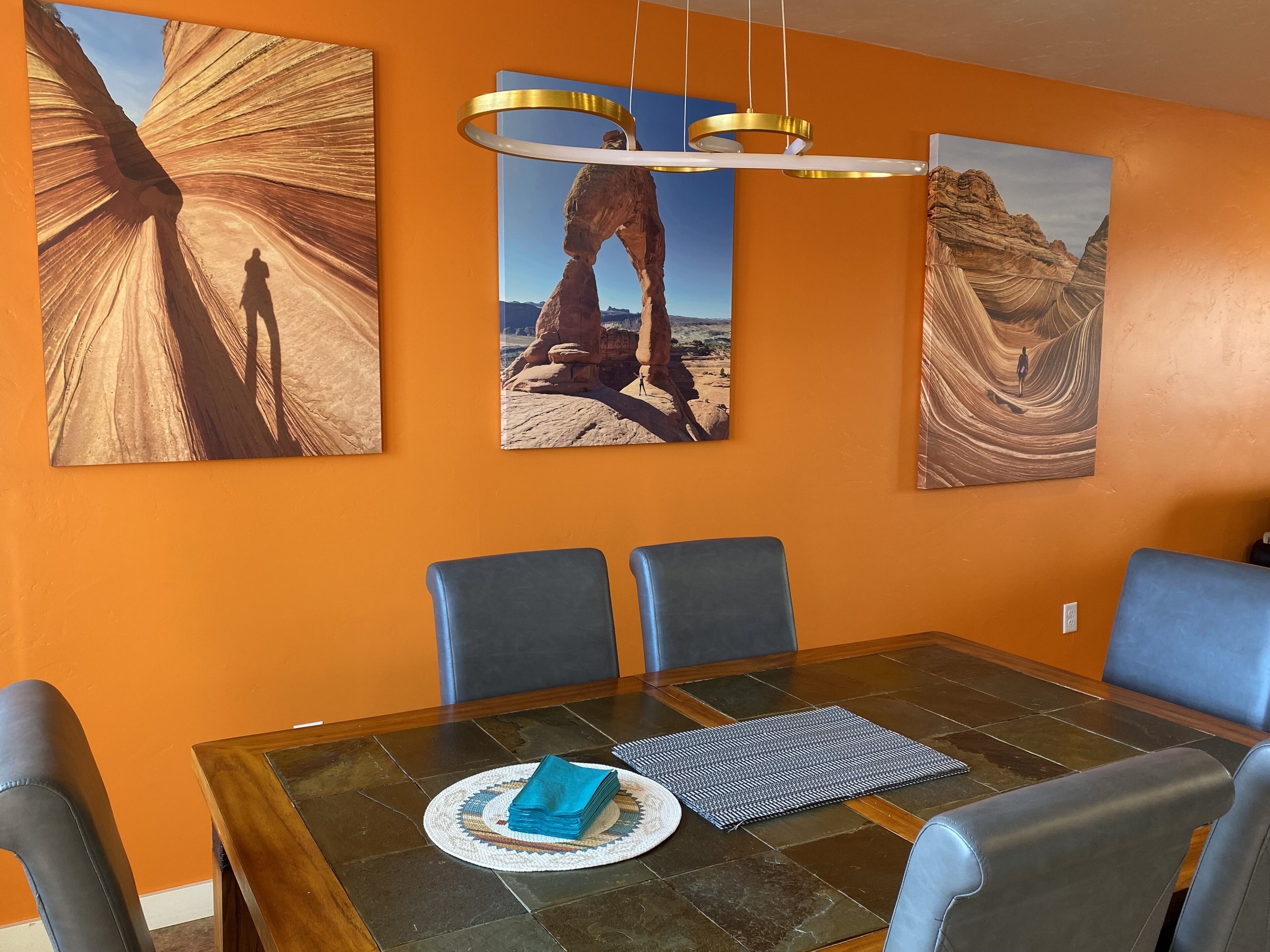 A dining area with a glass table, blue chairs, and three paintings of desert landscapes.