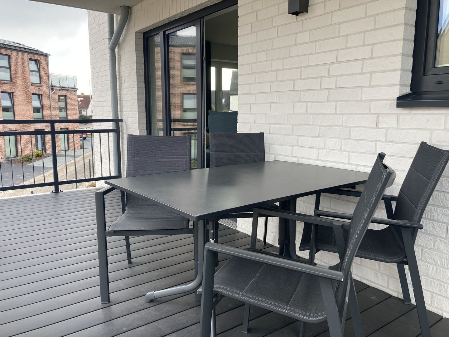 Un balcon avec une table et des chaises, une vue sur un immeuble en briques et une fenêtre avec des stores.