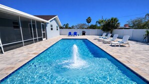 Una piscina rectangular con una fuente en el centro, rodeada de sillas de descanso y un área de terraza cubierta.