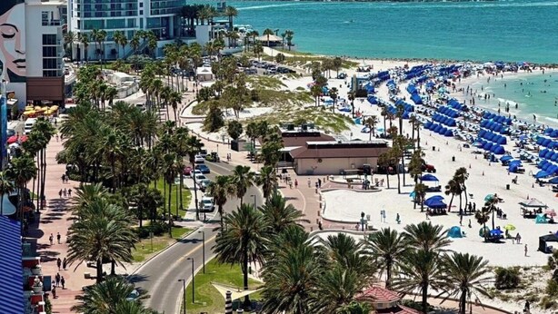 Vista marítima con un paseo bordeado de palmeras, una playa arenosa con sombrillas azules y rascacielos al fondo.