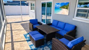Un patio techado con mobiliario de mimbre, un tapete azul con motivos y un cuadro con temática marina en la pared.