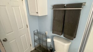 Un baño con inodoro blanco, un mueble blanco, una estantería metálica con dos toallas, un tendedero metálico y un bote de basura.