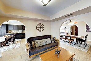 A spacious living area with a dark brown sofa, wooden coffee table, and a dining table with benches. A kitchen is visible through an archway, and a ceiling fan is present.
