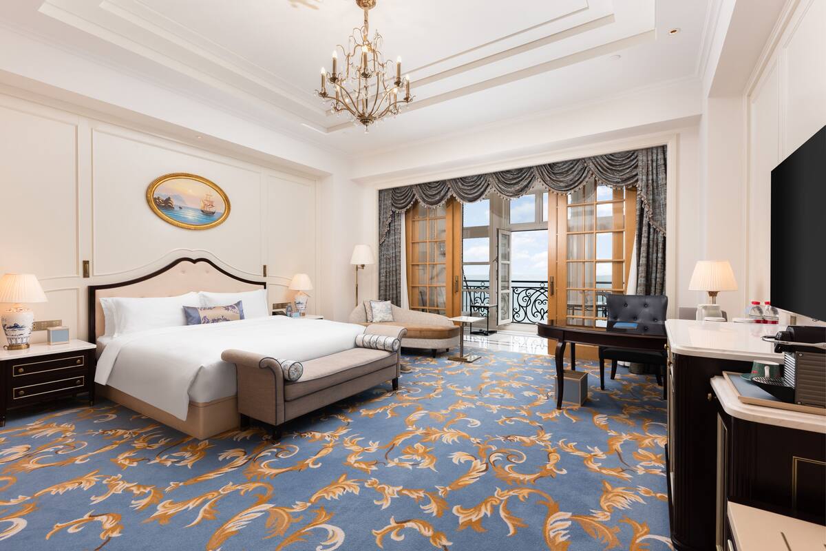 A spacious hotel room with a large bed, a sitting area with a sofa and armchairs, a dining table, and a view of the outdoors through a large window.