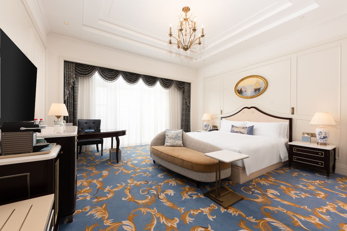 A hotel room with a large bed, a desk, a chair, a small table, and a chandelier.