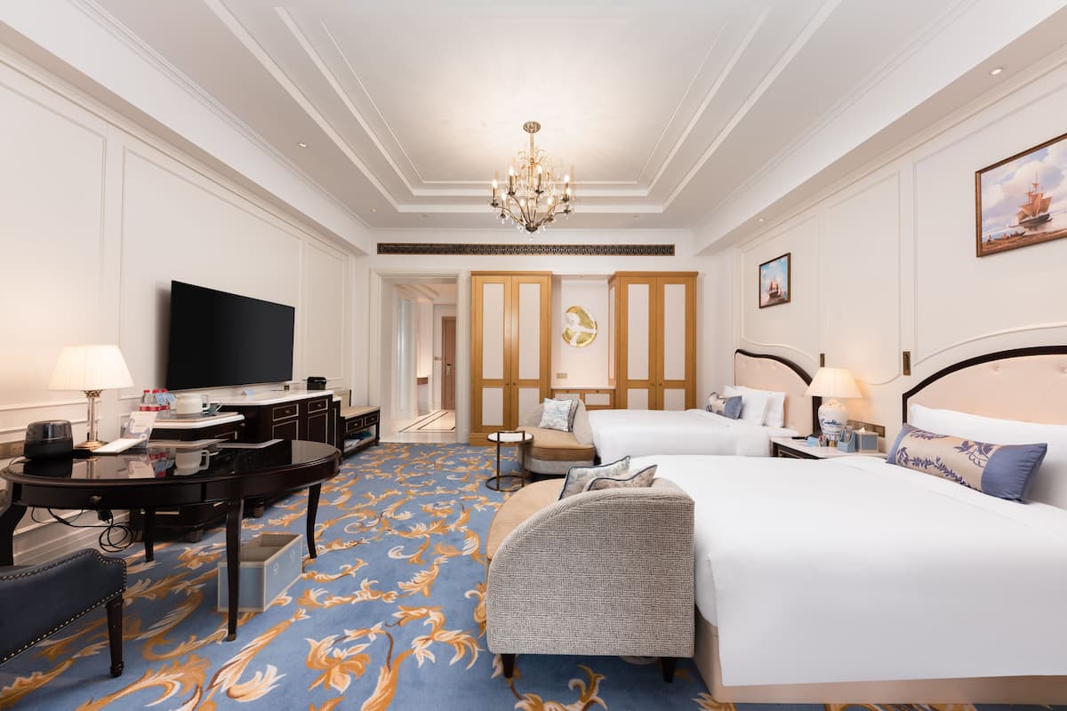 A hotel room with a large bed, a smaller bed, a desk with a flat-screen TV, a chair, and a decorative carpet.