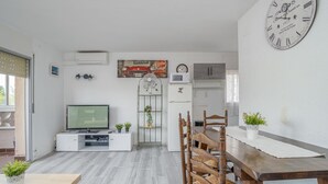 Un salon moderne avec un téléviseur, une table à manger avec des chaises, une horloge, et un coin cuisine en arrière-plan.