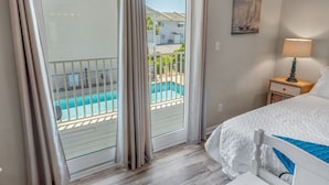 A bedroom with a bed, a nightstand with a lamp, a painting, and a view of a swimming pool and buildings through a sliding glass door. - Beachy Charm 2BR | Walk to Beach + Parking (Miramar Beach)