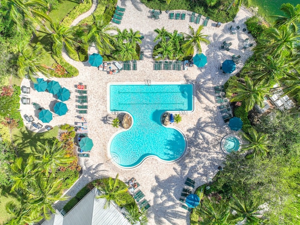 Vista aérea de un complejo turístico con una piscina central, rodeado de zonas de descanso con sombrillas y sillas, y palmeras.