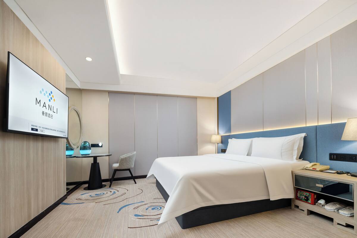 Ein modernes Hotelzimmer mit Bett, Nachttisch, Lampe und Fernseher, der den Namen des Hotels zeigt.