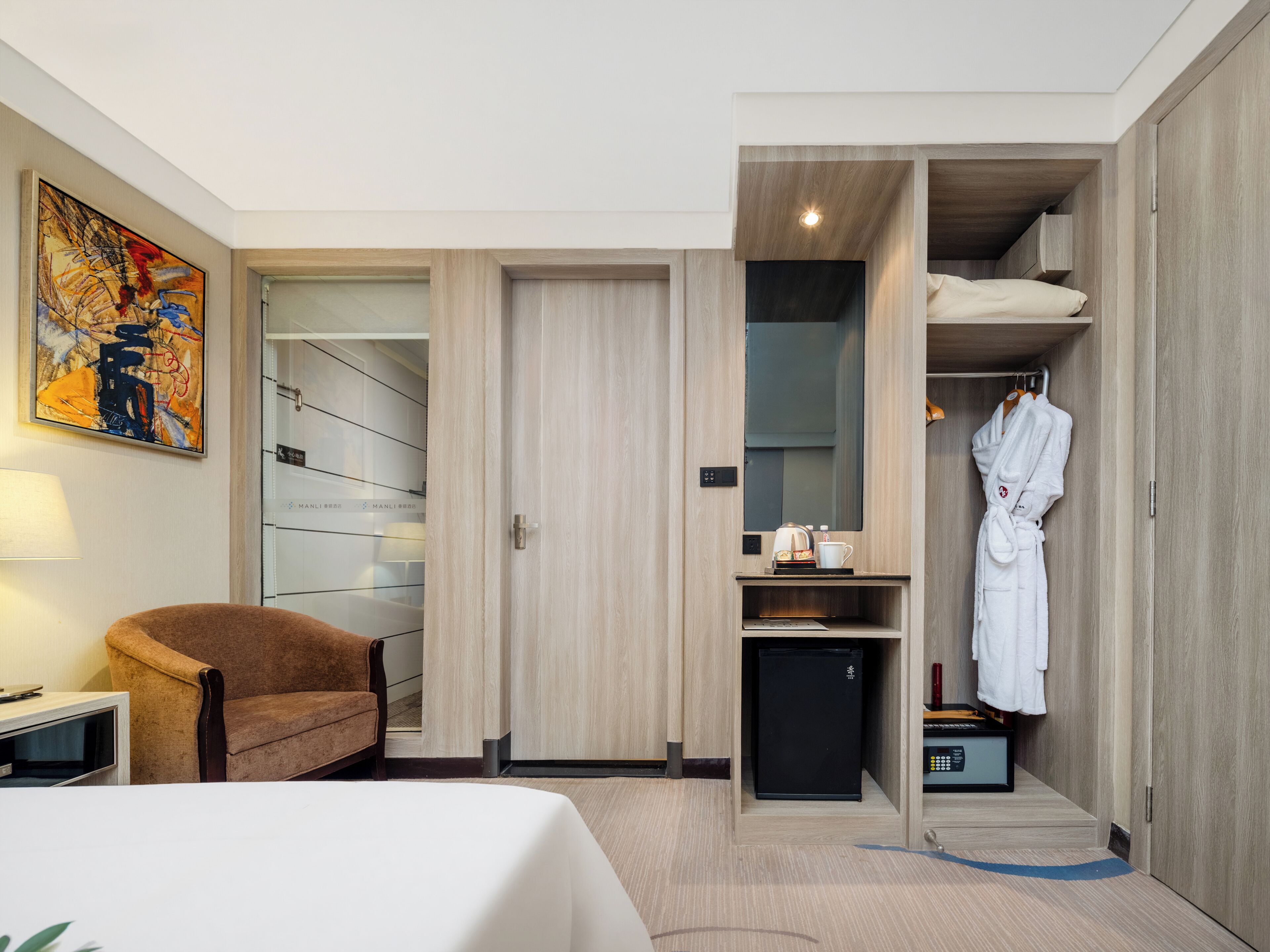 A hotel room with a bed, a chair, a wardrobe, a small fridge, and a painting on the wall.