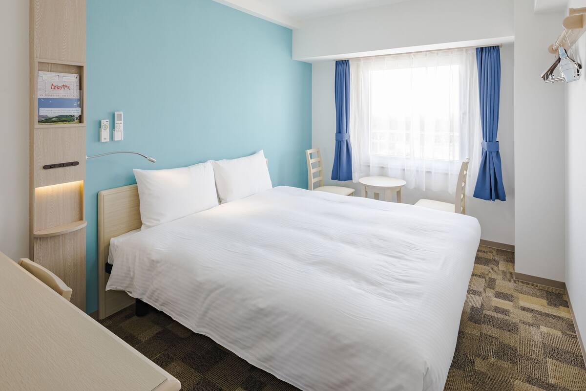 A hotel room with a large bed, a desk, and a chair. There is a window with blue curtains and a view of the outside.