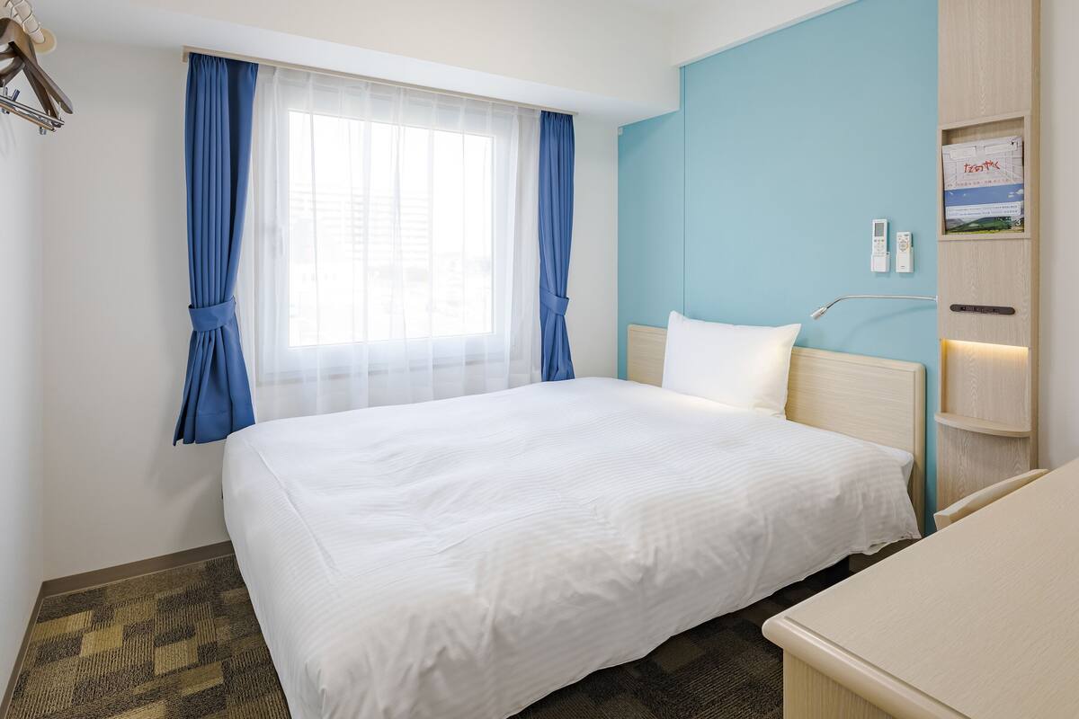 A hotel room with a large bed, a bedside table, a desk, a window with curtains, and a wall-mounted control panel.