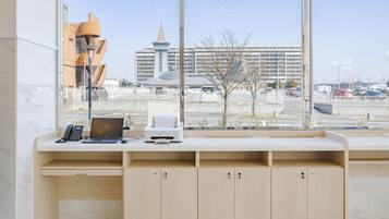 A modern office desk with a laptop, phone, and printer, large windows with a city view, and a clean, minimalist design.