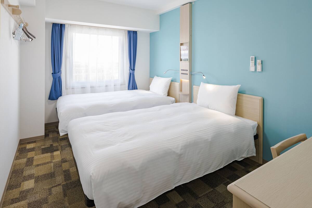 A hotel room with two beds, a desk, and a large window with curtains.