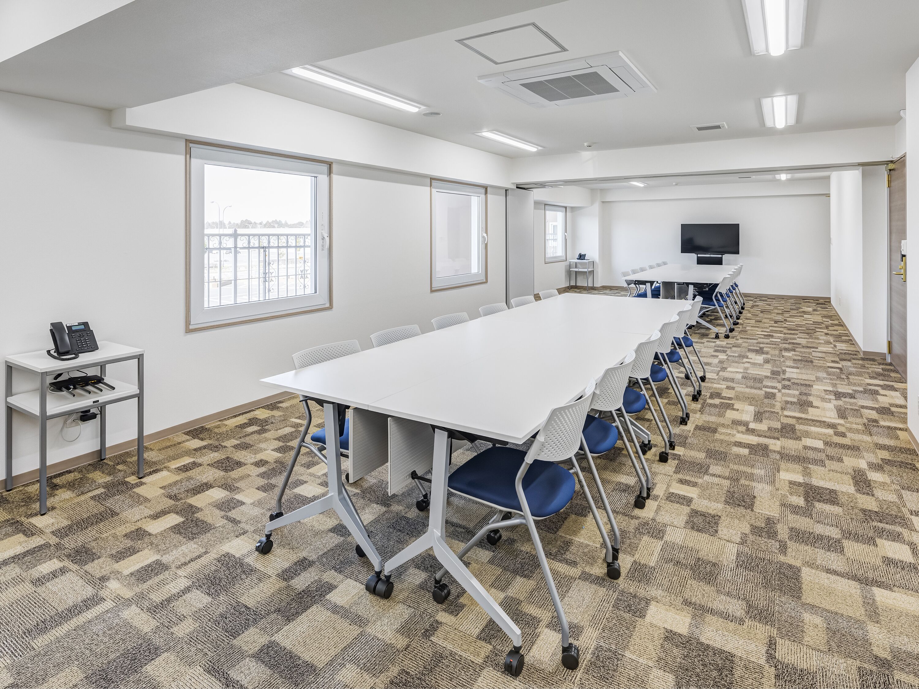 A conference room with a long table, chairs, a phone on a desk, and a TV on the wall.