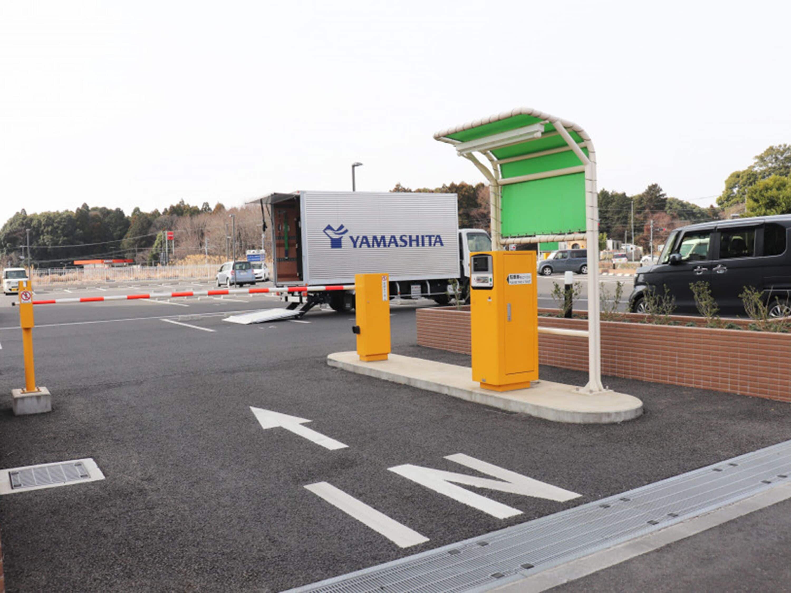 Un espace de stationnement où se trouve un camion blanc portant l’inscription « YAMASHITA » et une structure en forme de tonnelle, de couleur verte.