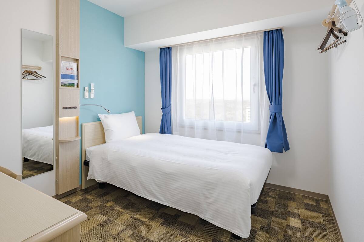 A hotel room with a bed, a bedside table, a mirror, a window with blue curtains, and a wall-mounted light fixture.