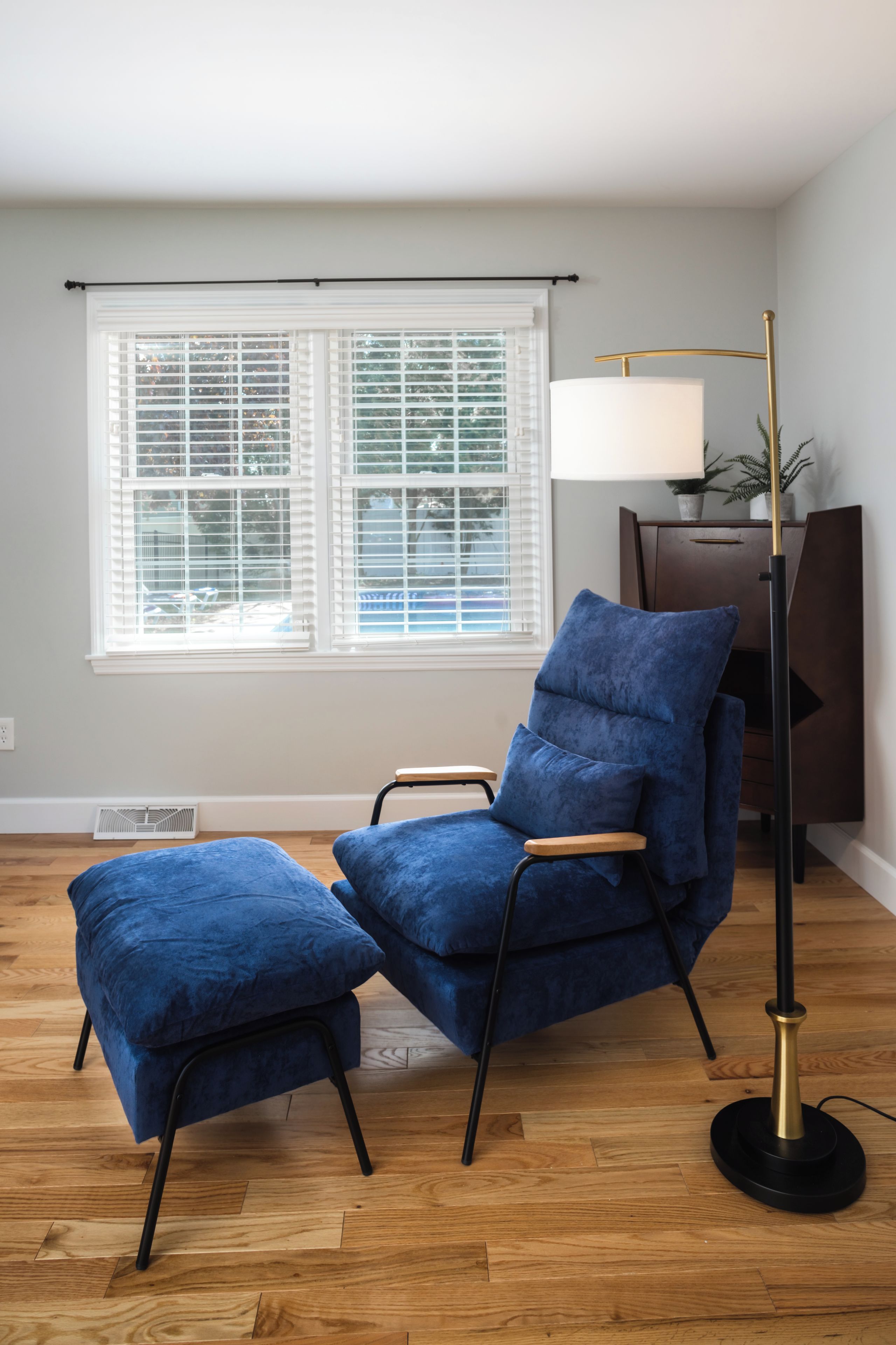 A room with a blue velvet armchair and ottoman, a floor lamp, and a window with blinds.