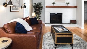 Ein Wohnzimmer mit braunem Ledersofa, einem runden Holztisch, einem Kamin und zwei Wandleuchten.