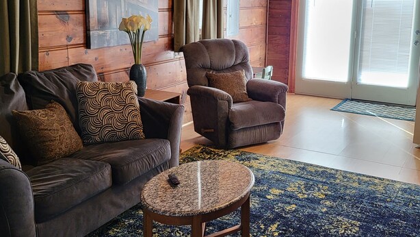 A cozy living room with a brown leather sofa, a brown armchair, a small round table, and a blue patterned rug. - Park Place-Quaint home nestled between parks and lakes (Olney)