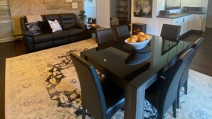 En moderne stue med en sort lædersofa, et spisebord med stole og et køkken i baggrunden.