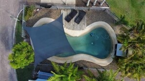 Ovenudblik over en swimmingpool med et mørkt skyggesejl, omgivet af palmer og en træterrasse.
