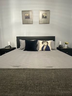 Uma cama bem arrumada com uma cabeceira escura, duas mesas de cabeceira com luminárias e duas imagens emolduradas na parede.
