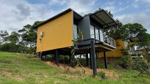 Una casa moderna de dos pisos con fachada amarilla, elevada sobre pilotes, que cuenta con un balcón y barandilla de metal.