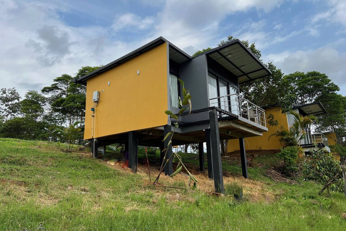 A modern two-story house with a yellow exterior, elevated on stilts, featuring a balcony with a metal railing.