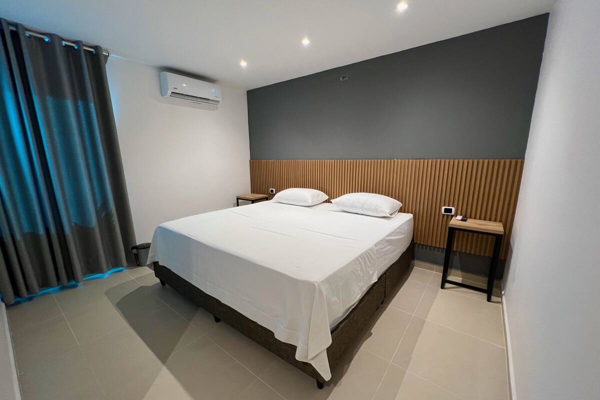 A modern hotel room with a large bed, a bedside table, an air conditioning unit, and a curtain.