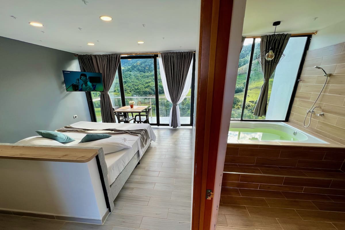 A modern bedroom with a bed, a small table with chairs, a TV, and a large window with curtains.