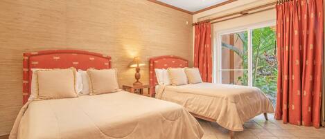 A hotel room with two beds, a ceiling fan, a window with red curtains, and a view of greenery outside.