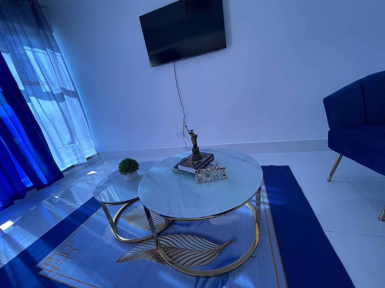 Una sala de estar moderna con una mesa de centro de vidrio, un sofá azul y un televisor de pantalla plana montado en la pared.