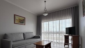 Sala de estar com sofá cinza, mesa de centro de madeira, TV sobre um suporte de madeira e varanda com vista.